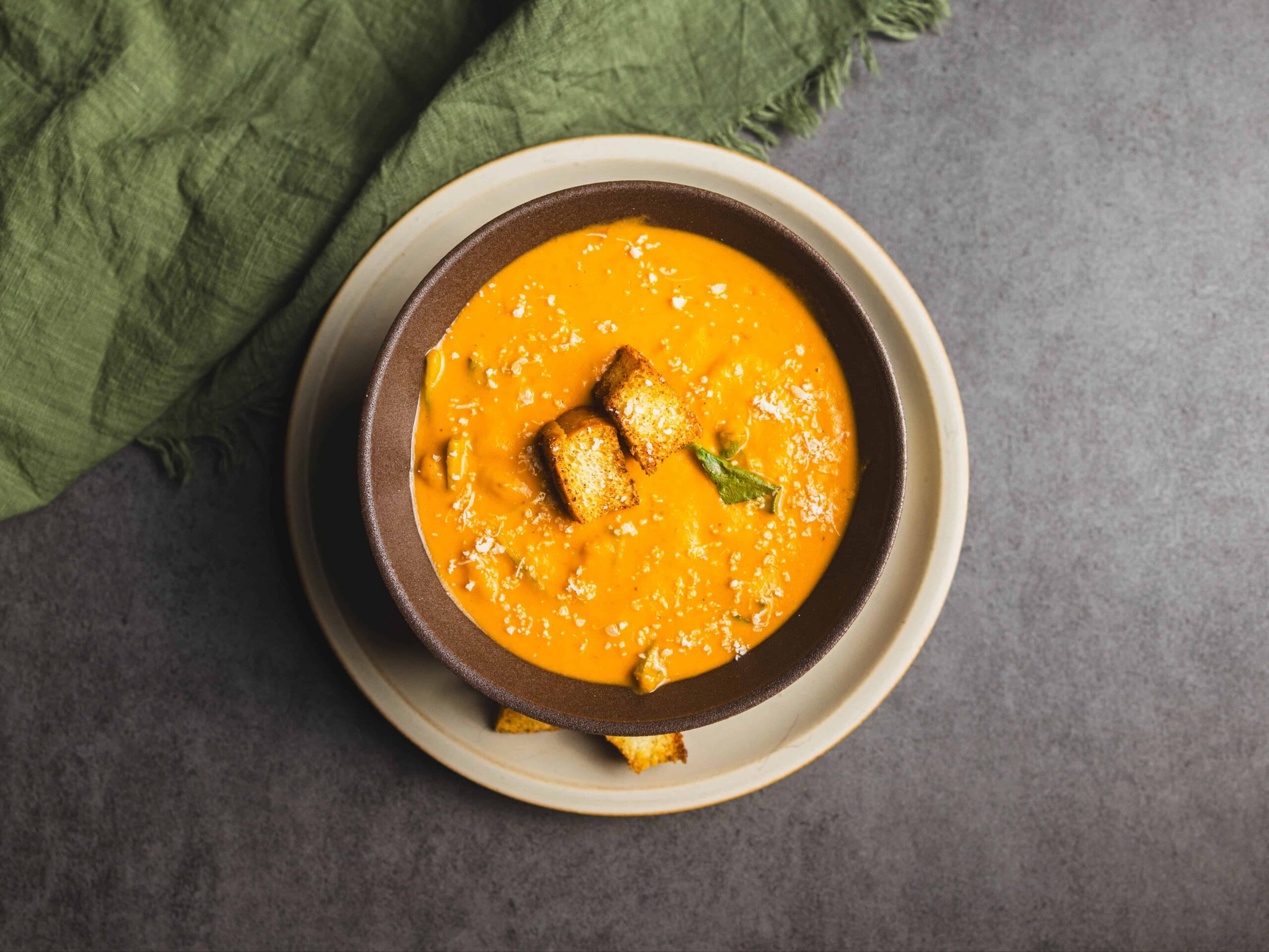 Creamy Butternut Squash Soup garnished with croutons and Parmesan cheese, served in a brown bowl on a beige plate, with a green napkin on a dark grey surface.