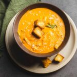 Creamy butternut squash soup garnished with croutons and a fresh sage leaf on top, served in a rustic brown bowl. Perfect for fall comfort food and healthy eating.