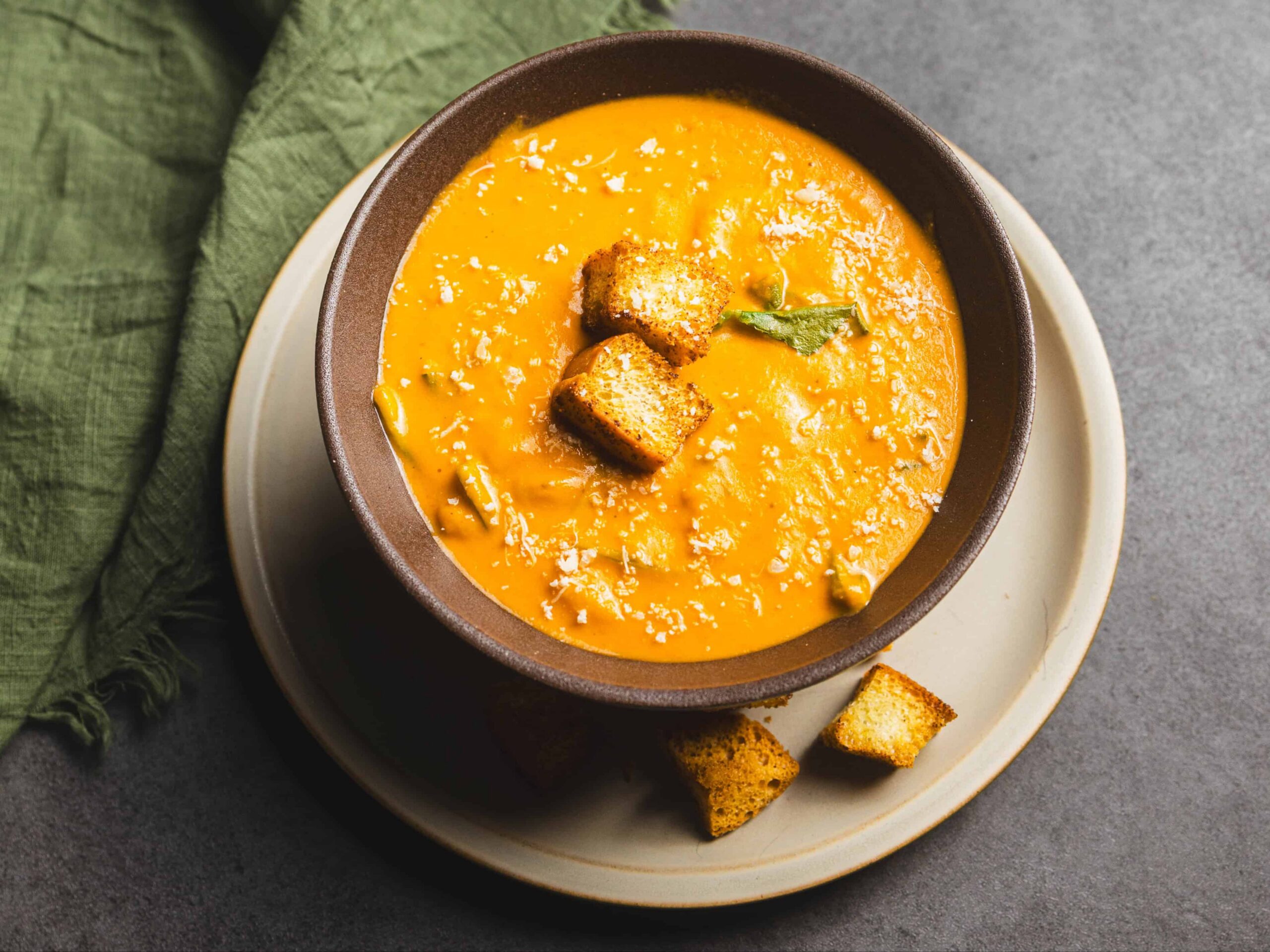 Creamy butternut squash soup garnished with croutons and a fresh sage leaf on top, served in a rustic brown bowl. Perfect for fall comfort food and healthy eating.