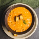 Creamy butternut squash soup garnished with croutons and fresh herbs, served in a bowl on a rustic table with green cloth napkin, perfect for cozy fall meals.
