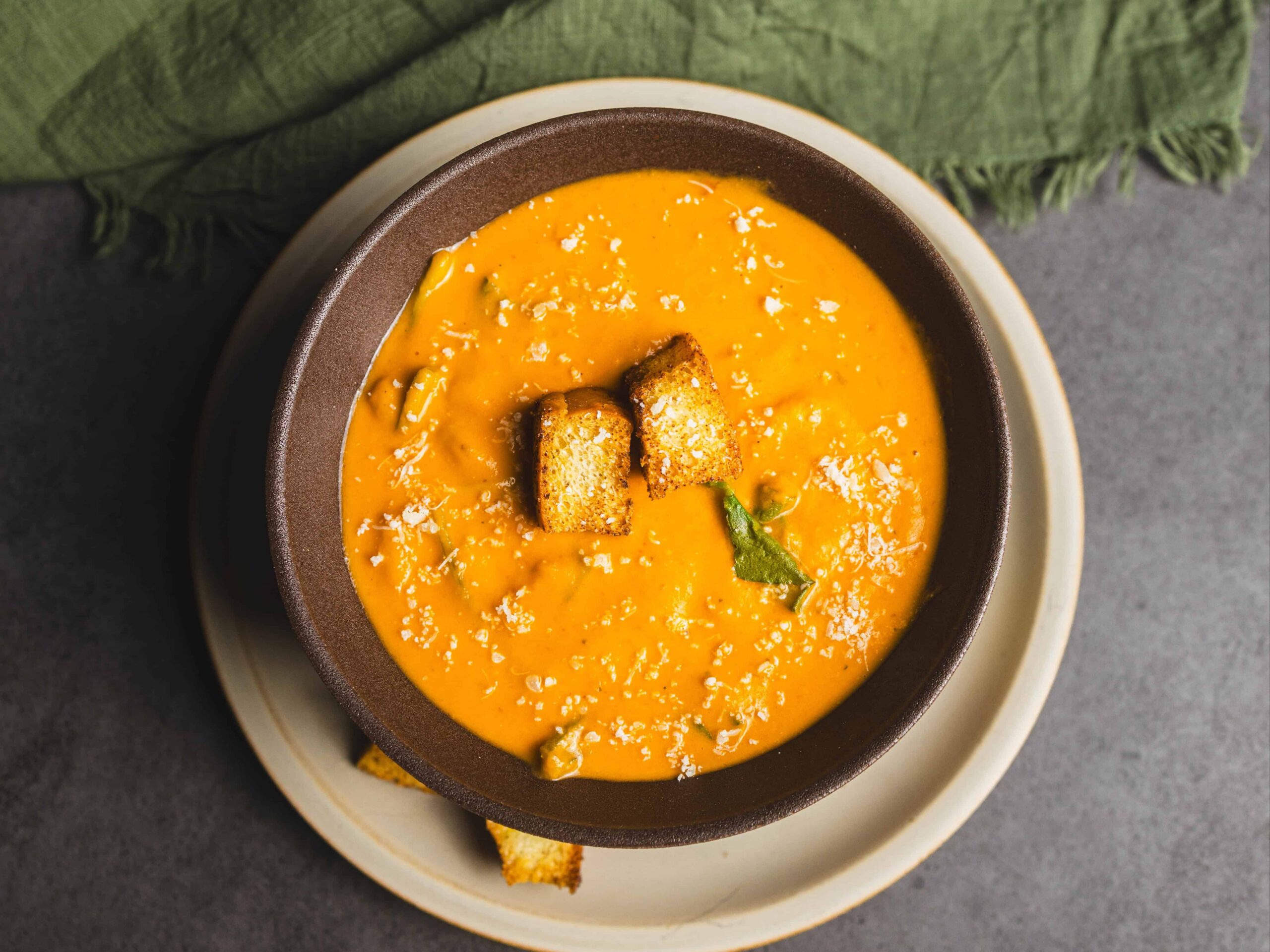 Creamy butternut squash soup garnished with croutons and fresh herbs, served in a bowl on a rustic table with green cloth napkin, perfect for cozy fall meals.